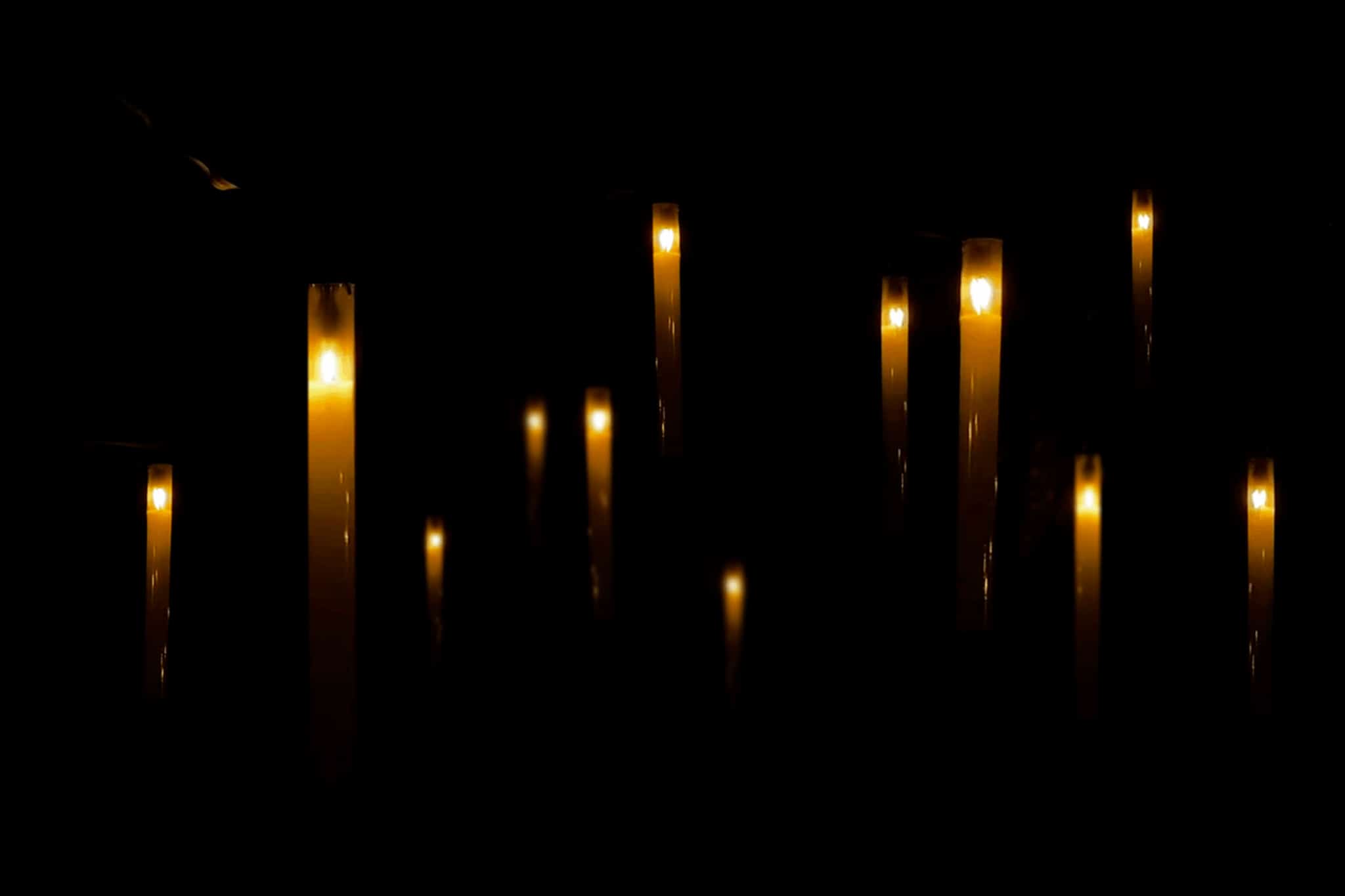 Plusieurs bougies allumées brillent doucement dans l'obscurité, leur lumière se reflétant sur des surfaces brillantes. La scène est faiblement éclairée, créant une atmosphère calme et sereine.