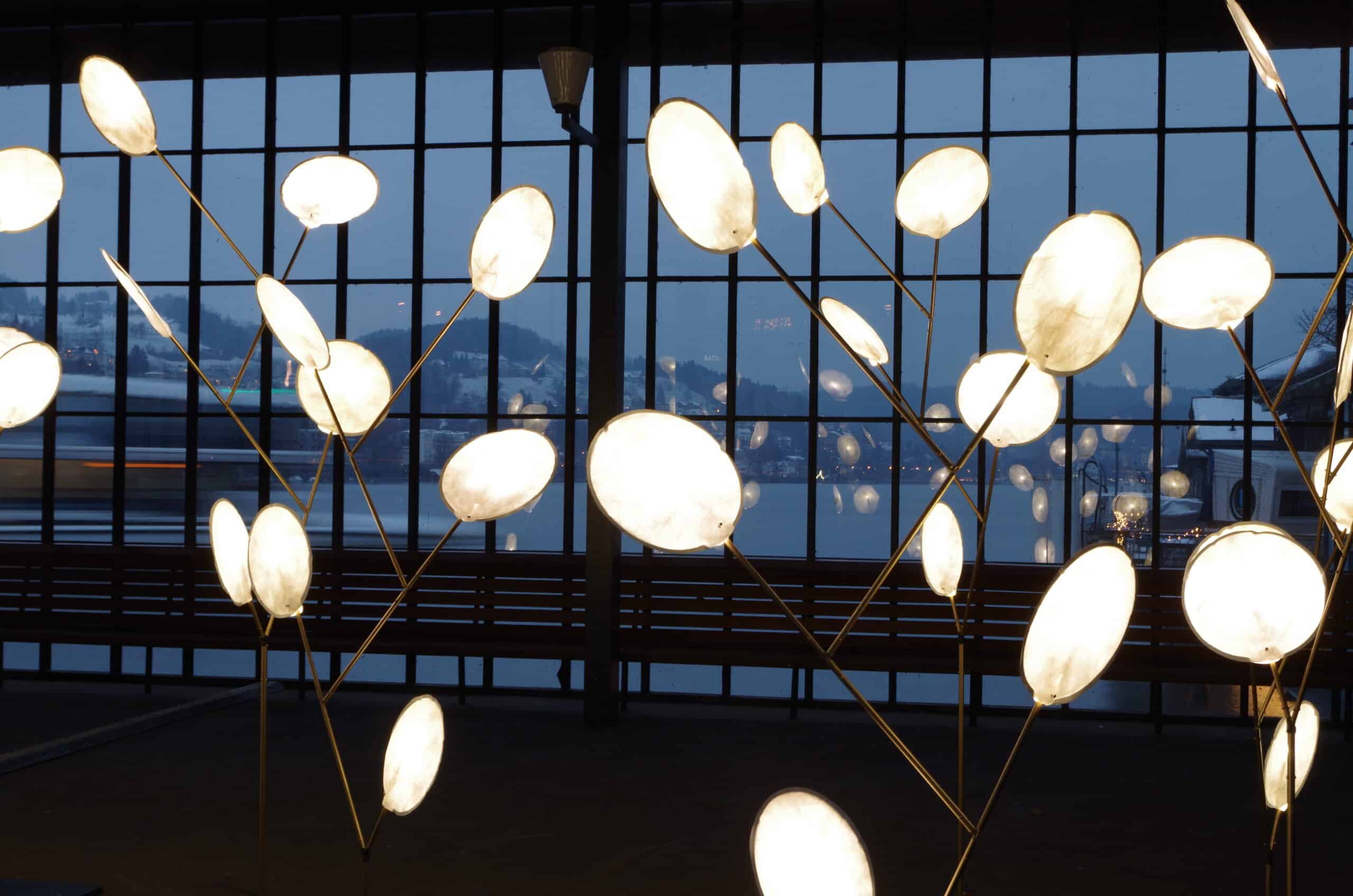 Installation artistique composée de lumières circulaires brillantes montées sur de minces poteaux, placées contre de grandes fenêtres offrant une vue sur un lac et des collines au crépuscule. Les lumières ressemblent à des nénuphars illuminés ou à des fleurs abstraites.