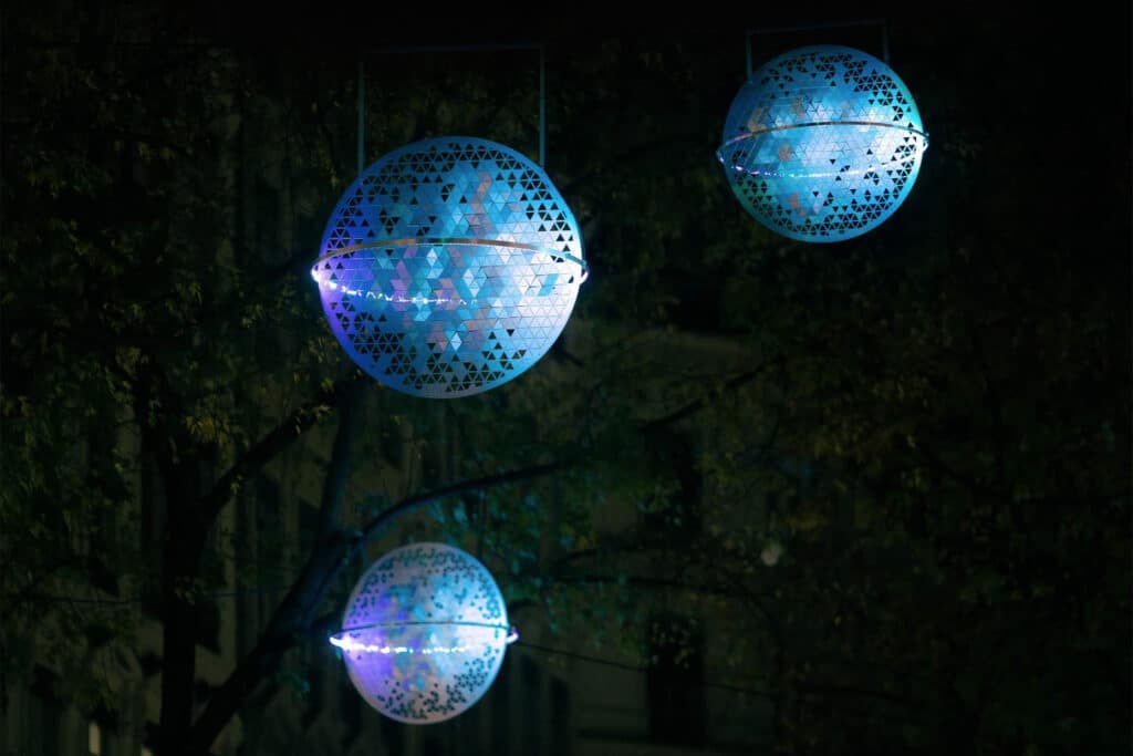 Trois installations lumineuses sphériques aux teintes bleues et violettes sont suspendues à l'extérieur la nuit, entourées d'arbres et d'obscurité, créant une atmosphère éthérée.