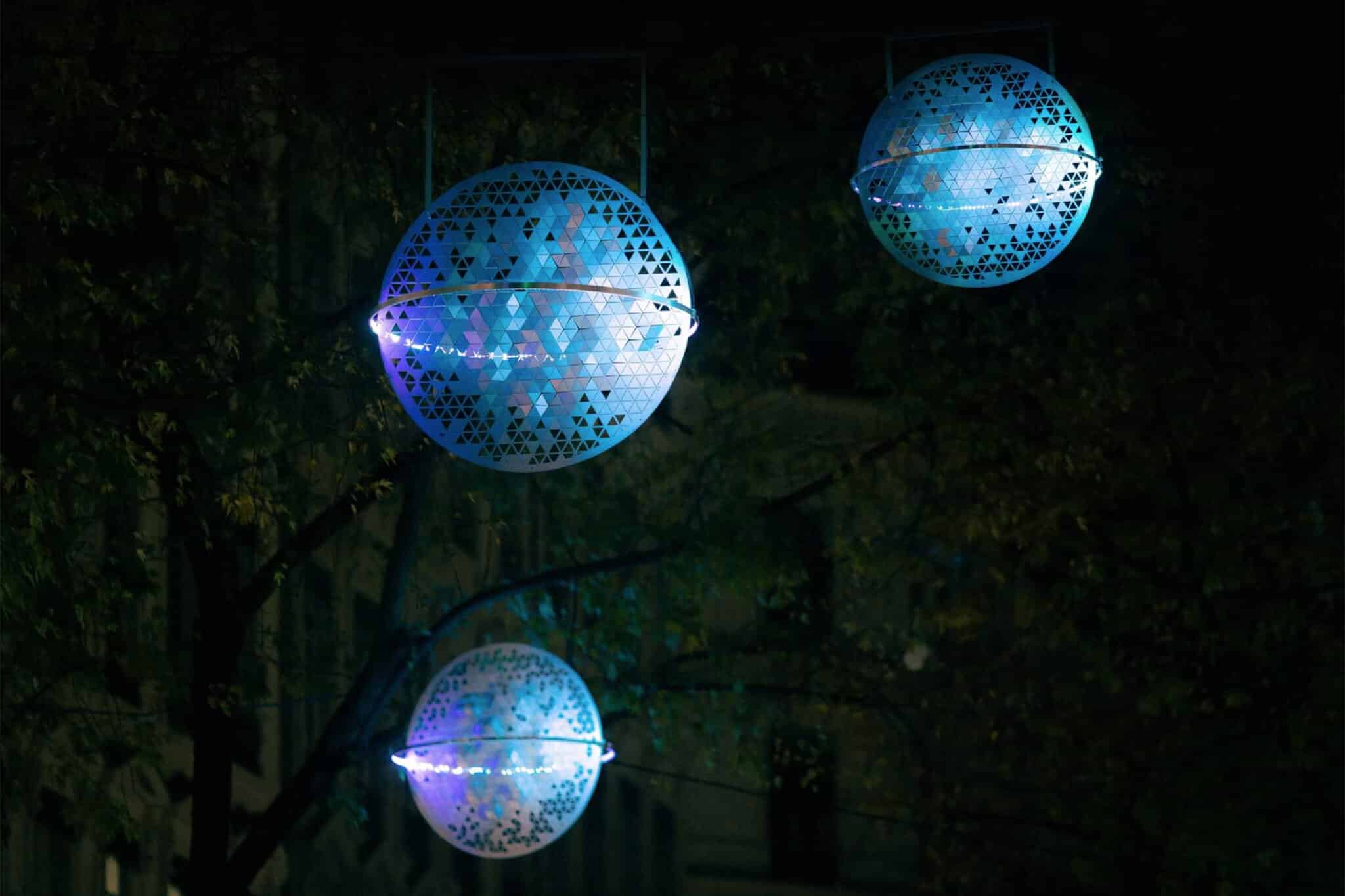 Trois installations lumineuses sphériques aux teintes bleues et violettes sont suspendues à l'extérieur la nuit, entourées d'arbres et d'obscurité, créant une atmosphère éthérée.