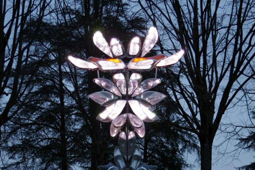 Une sculpture composée de pièces métalliques illuminées en forme de pétales s'illumine de lumières blanches et orange devant des arbres dépourvus de feuilles sur fond de ciel crépusculaire.
