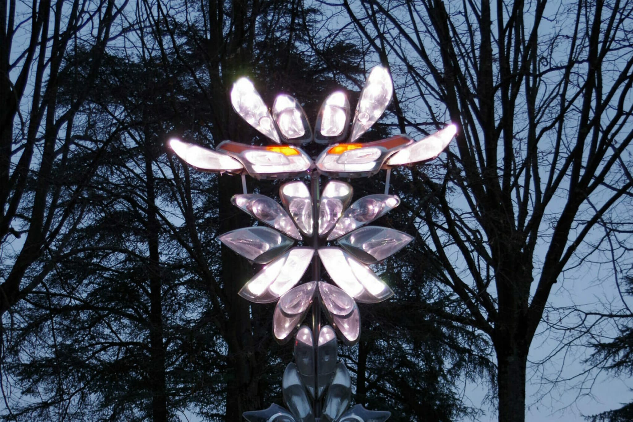 Une sculpture composée de pièces métalliques illuminées en forme de pétales s'illumine de lumières blanches et orange devant des arbres dépourvus de feuilles sur fond de ciel crépusculaire.