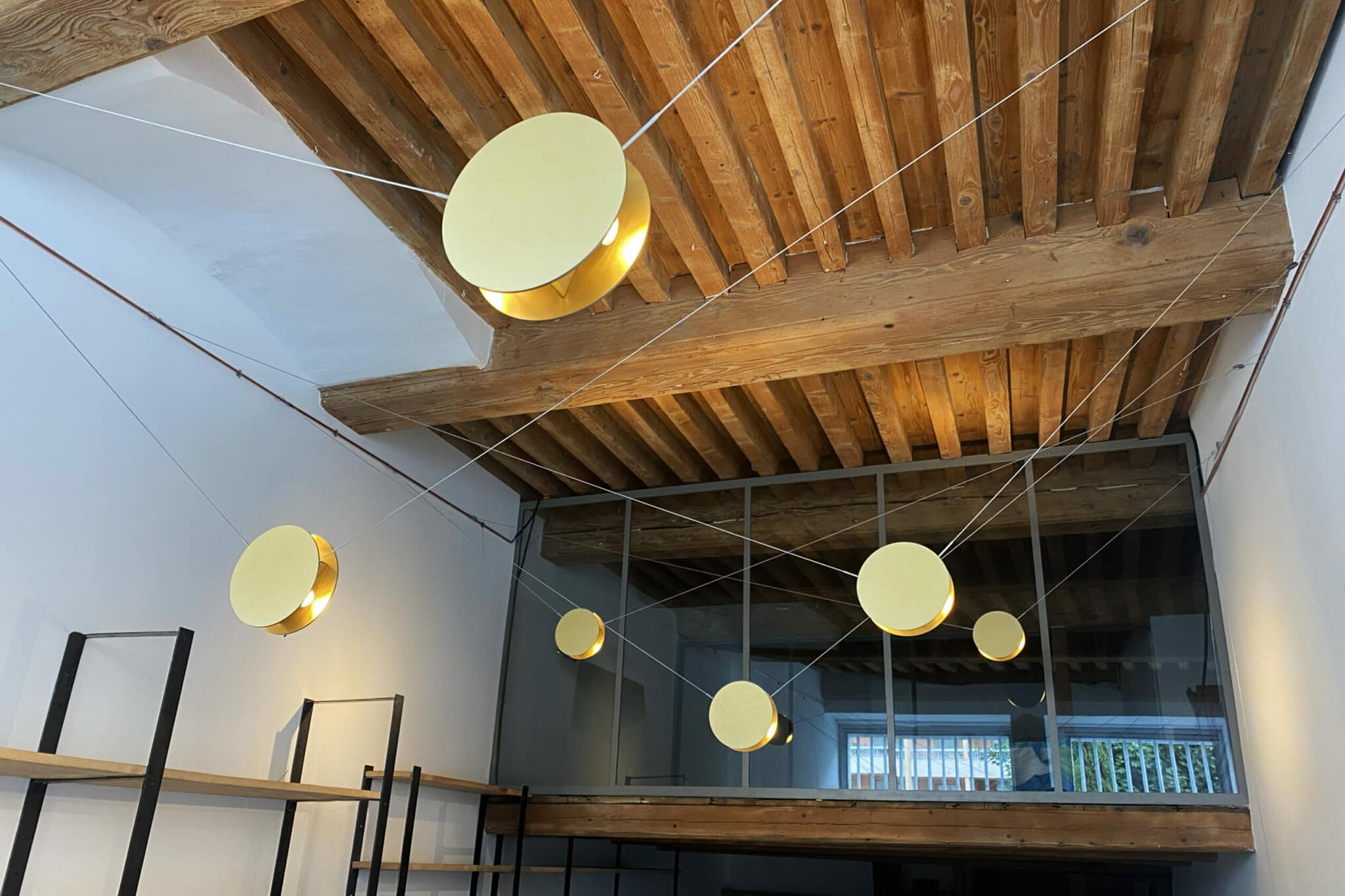Un intérieur moderne présente un plafond à poutres en bois avec des lampes suspendues rondes et dorées à différentes hauteurs, suspendues par des cordons blancs. Au-dessous se trouve une mezzanine et des étagères en métal noir.